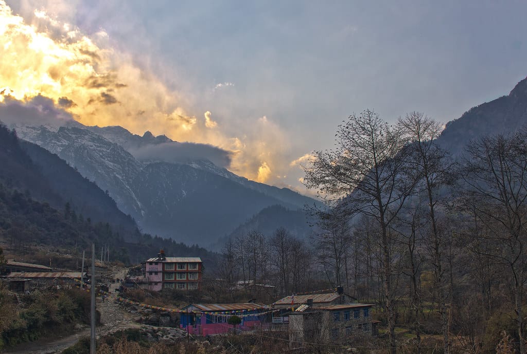 Trailside lodges in a Himalayan valley at dusk, snow peaks under golden cloud